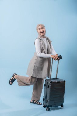 Gri yelek ve ten rengi pantolon giyen bir kadın gülümsüyor ve elinde bavul tutuyor. Çanta siyah ve tekerlekleri var.