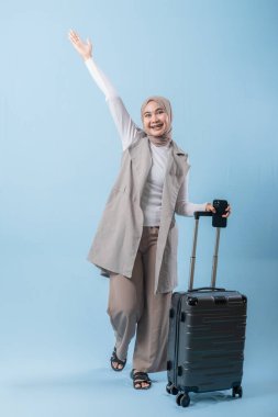 Bir kadın bir bavul tutuyor ve gülümsüyor. Beyaz bir gömlek ve gri bir yelek giyiyor.