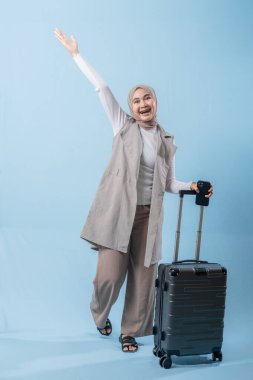 Bir kadın bir bavul tutuyor ve gülümsüyor. Beyaz bir gömlek ve gri bir yelek giyiyor.