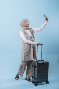 Eşarp takan ve cep telefonu tutan bir kadın bir bavulun yanında duruyor. Bavulla kendi fotoğrafını çekiyor.