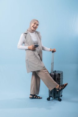 Eşarp takan ve cep telefonu tutan bir kadın bir bavulun yanında duruyor. O, yaklaşan gezisi hakkında mutlu ve heyecanlı.