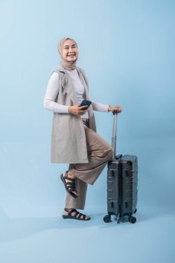 Başörtüsü takan bir kadın bir bavul ve cep telefonuyla yürüyor. O mutlu ve seyahatinin tadını çıkarıyor.