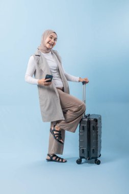 Başörtüsü takan bir kadın bir bavul ve cep telefonuyla yürüyor. O mutlu ve seyahatinin tadını çıkarıyor.