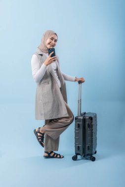 Eşarp takan ve cep telefonu tutan bir kadın bir bavulun yanında duruyor. Bavulla kendi fotoğrafını çekiyor.