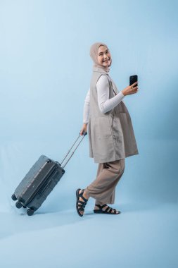 Başörtüsü takan bir kadın bir bavul ve cep telefonuyla yürüyor. O mutlu ve seyahatinin tadını çıkarıyor.