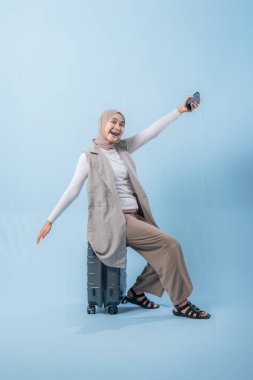 Başörtüsü takan bir kadın bavulla poz veriyor. Gülümsüyor ve mutlu.