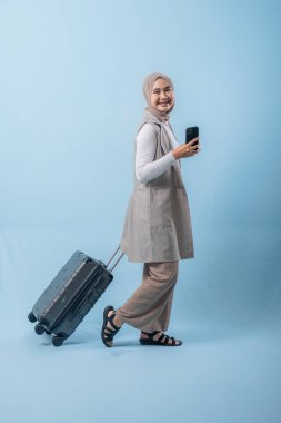 Başörtüsü takan bir kadın bir bavul ve cep telefonuyla yürüyor. O mutlu ve seyahatinin tadını çıkarıyor.