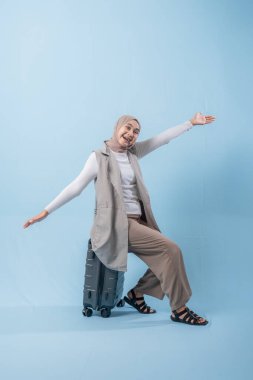 Başörtüsü takan bir kadın bavulla poz veriyor. Gülümsüyor ve mutlu.