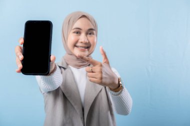 Bir kadın elinde siyah bir cep telefonu tutuyor ve onu işaret ediyor. Gülümsüyor ve mutlu.