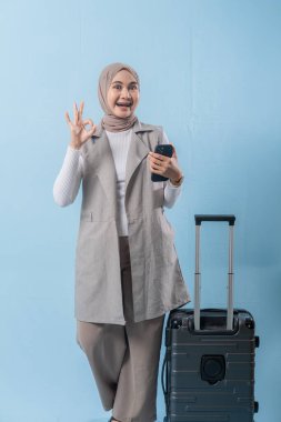 Başörtüsü takan bir kadın cep telefonu tutuyor ve gülümsüyor. Bir bavulun yanında duruyor.