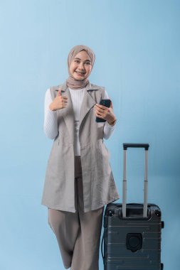 Başörtüsü takan bir kadın cep telefonu tutuyor ve gülümsüyor. Bir bavulun yanında duruyor.