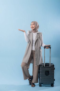 Beyaz gömlek ve gri yelek giyen bir kadın bir bavul tutuyor ve önemli bir şeye işaret ediyor.