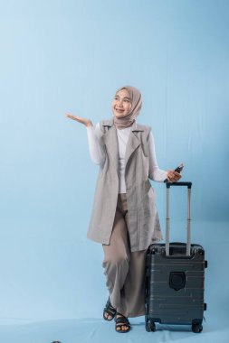 Beyaz gömlek ve gri yelek giyen bir kadın bir bavul tutuyor ve önemli bir şeye işaret ediyor.