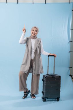 Beyaz gömlek ve gri yelek giyen bir kadın bir bavul tutuyor ve önemli bir şeye işaret ediyor.