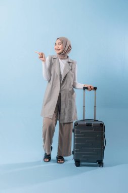 Beyaz gömlek ve gri yelek giyen bir kadın bir bavul tutuyor ve önemli bir şeye işaret ediyor.