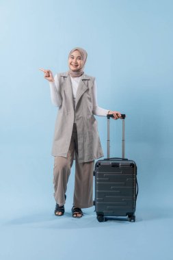 Beyaz gömlek ve gri yelek giyen bir kadın bir bavul tutuyor ve önemli bir şeye işaret ediyor.