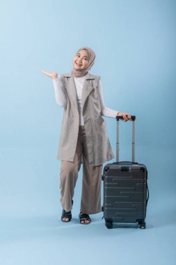 Beyaz gömlek ve gri yelek giyen bir kadın bir bavul tutuyor ve önemli bir şeye işaret ediyor.