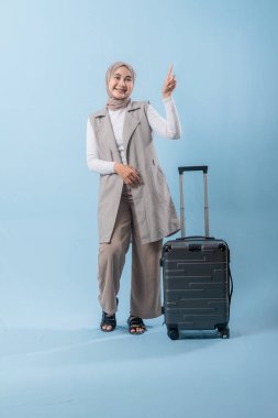 Beyaz gömlek ve gri yelek giyen bir kadın bir bavul tutuyor ve önemli bir şeye işaret ediyor.