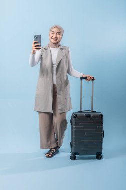 Bir kadın elinde bavul tutarken telefonuyla selfie çekiyor. Seyahat ve macera kavramı, kadın bir yolculuğa çıkmaya hazırlanırken.