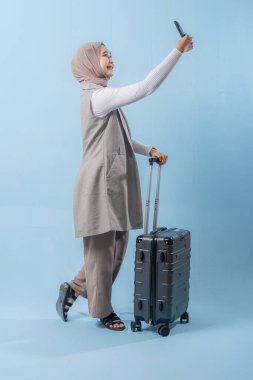 Bir kadın elinde bavul tutarken telefonuyla selfie çekiyor. Seyahat ve macera kavramı, kadın bir yolculuğa çıkmaya hazırlanırken.