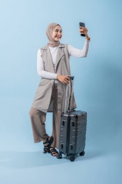 Bir kadın elinde bavul tutarken telefonuyla selfie çekiyor. Seyahat ve macera kavramı, kadın bir yolculuğa çıkmaya hazırlanırken.