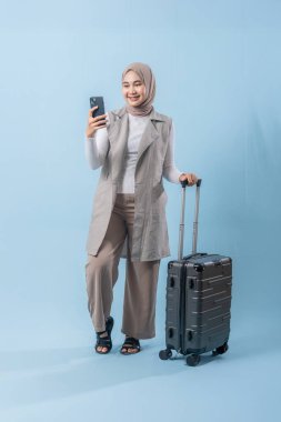 Başörtüsü takan bir kadın cep telefonu tutuyor ve gülümsüyor. Bir bavulun yanında duruyor.