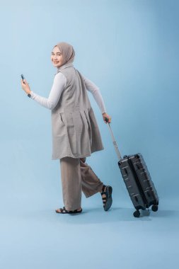 Başörtüsü takan bir kadın bir bavul ve cep telefonuyla yürüyor. O mutlu ve seyahatinin tadını çıkarıyor.