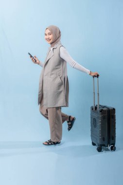 Başörtüsü takan bir kadın bir bavul ve cep telefonuyla yürüyor. O mutlu ve seyahatinin tadını çıkarıyor.