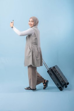 Başörtüsü takan bir kadın bir bavul ve cep telefonuyla yürüyor. O mutlu ve seyahatinin tadını çıkarıyor.