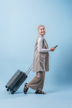 Başörtüsü takan bir kadın bir bavul ve cep telefonuyla yürüyor. O mutlu ve seyahatinin tadını çıkarıyor.