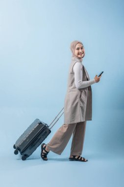 Başörtüsü takan bir kadın bir bavul ve cep telefonuyla yürüyor. O mutlu ve seyahatinin tadını çıkarıyor.