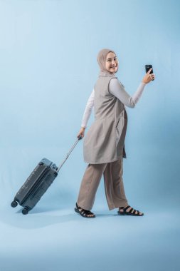 Başörtüsü takan bir kadın bir bavul ve cep telefonuyla yürüyor. O mutlu ve seyahatinin tadını çıkarıyor.