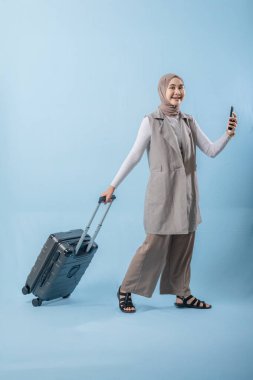 Başörtüsü takan bir kadın bir bavul ve cep telefonuyla yürüyor. O mutlu ve seyahatinin tadını çıkarıyor.