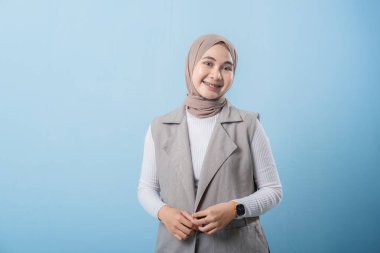 Mavi bir duvarın önünde beyaz gömlek ve bronz bir eşarp giyen bir kadın duruyor. Yüzünde bir gülümseme var ve mutlu.