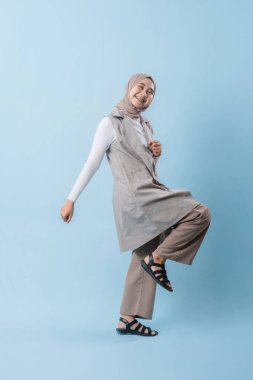 Gri yelek ve beyaz gömlek giyen bir kadın kameraya gülümsüyor. Başörtüsü takıyor ve kollarını kavuşturuyor.