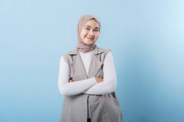 Gri yelek ve beyaz gömlek giyen bir kadın kameraya gülümsüyor. Başörtüsü takıyor ve kollarını kavuşturuyor.