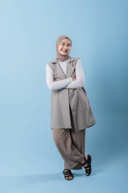 Gri yelek ve beyaz gömlek giyen bir kadın kameraya gülümsüyor. Başörtüsü takıyor ve kollarını kavuşturuyor.