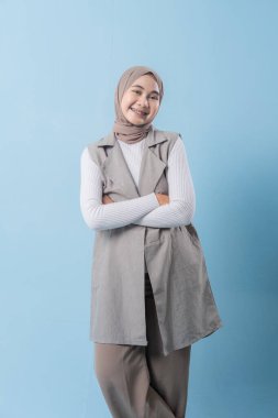 Gri yelek ve beyaz gömlek giyen bir kadın kameraya gülümsüyor. Başörtüsü takıyor ve kollarını kavuşturuyor.
