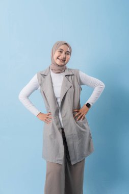 Gri yelek ve beyaz gömlek giyen bir kadın kameraya gülümsüyor. Başörtüsü takıyor ve kollarını kavuşturuyor.