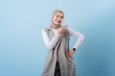 Gri yelek ve beyaz gömlek giyen bir kadın önemli bir şeyi işaret ediyor. Yüzünde bir gülümseme var ve bir türban takıyor.