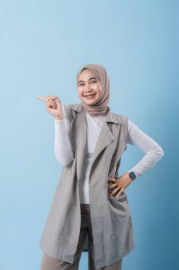 Gri yelek ve beyaz gömlek giyen bir kadın önemli bir şeyi işaret ediyor. Yüzünde bir gülümseme var ve bir türban takıyor.