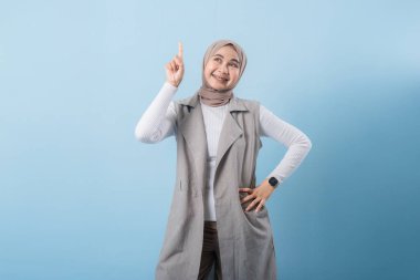 Gri yelek ve beyaz gömlek giyen bir kadın önemli bir şeyi işaret ediyor. Yüzünde bir gülümseme var ve bir türban takıyor.