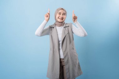 Gri yelek ve beyaz gömlek giyen bir kadın önemli bir şeyi işaret ediyor. Yüzünde bir gülümseme var ve bir türban takıyor.