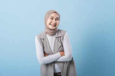 Gri yelek ve beyaz gömlek giyen bir kadın kameraya gülümsüyor. Başörtüsü takıyor ve kollarını kavuşturuyor.