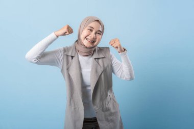 Gri yelekli bir kadın gülümsüyor ve kollarını kaldırıyor. Başörtüsü takıyor ve mutlu.