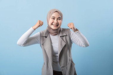 Gri yelekli bir kadın gülümsüyor ve kollarını kaldırıyor. Başörtüsü takıyor ve mutlu.