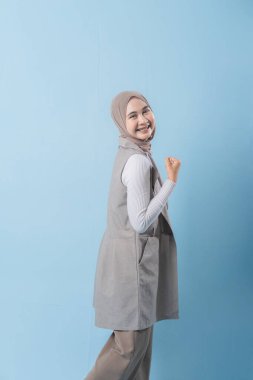 Gri yelekli bir kadın gülümsüyor ve kollarını kaldırıyor. Başörtüsü takıyor ve mutlu.