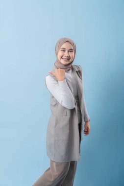 Gri yelekli bir kadın gülümsüyor ve kollarını kaldırıyor. Başörtüsü takıyor ve mutlu.