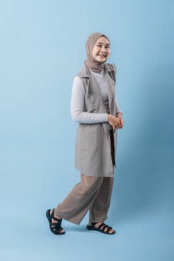 Gri yelek ve beyaz gömlek giyen bir kadın kameraya gülümsüyor. Başörtüsü takıyor ve kollarını kavuşturuyor.