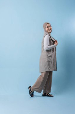 Gri yelek ve beyaz gömlek giyen bir kadın kameraya gülümsüyor. Başörtüsü takıyor ve kollarını kavuşturuyor.